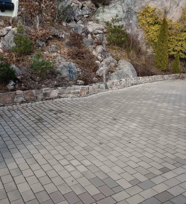 Tun lagt med grå belegningsstein, kantet med naturstein mot en fjellskrent.