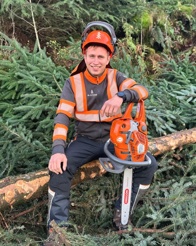 Vegard Bjerge sitter på en felt stamme i skogen, i vernetøy og hjelm, med motorsagen ved siden av seg.
